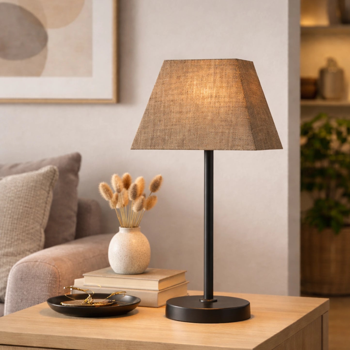 Quadratischer brauner Lampenschirm auf einer schlanken schwarzen Tischleuchte im Wohnzimmer; steht auf einem Holz‑Beistelltisch neben einer Vase mit Trockenblumen und einem Sofa; warmes Licht sorgt für behagliche Stimmung,Artikelnummer 30425-636.