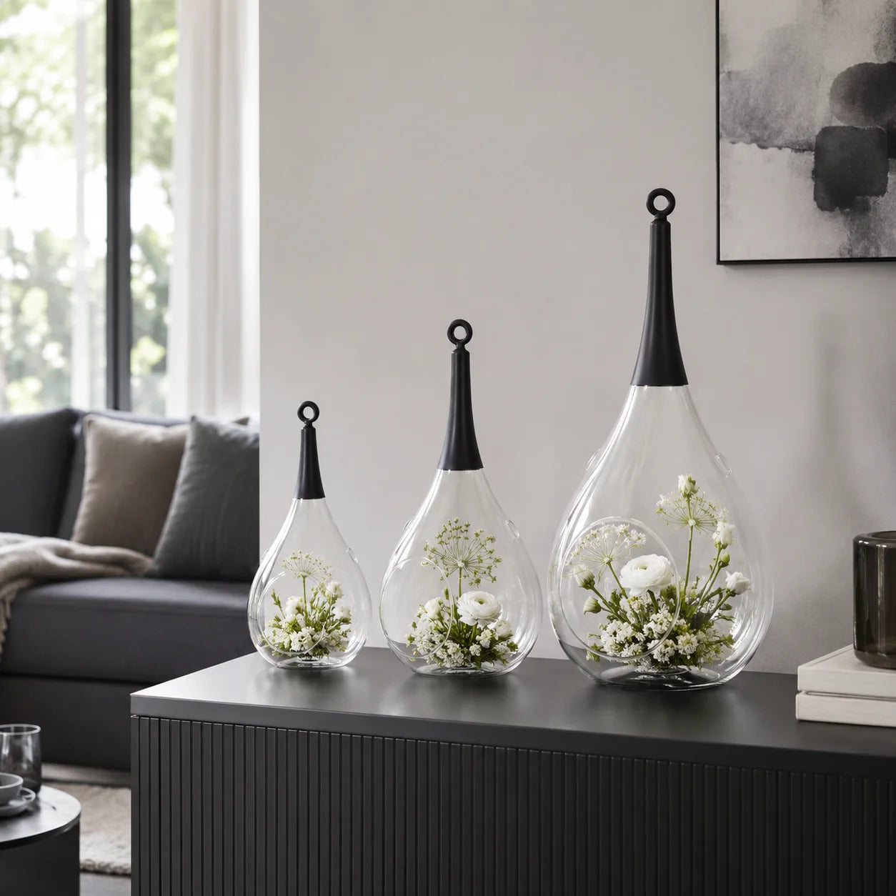 Drei Taura Glas-Windlichter in verschiedenen Größen mit weißen Blumen auf einem schwarzen Sideboard im modernen Wohnzimmer