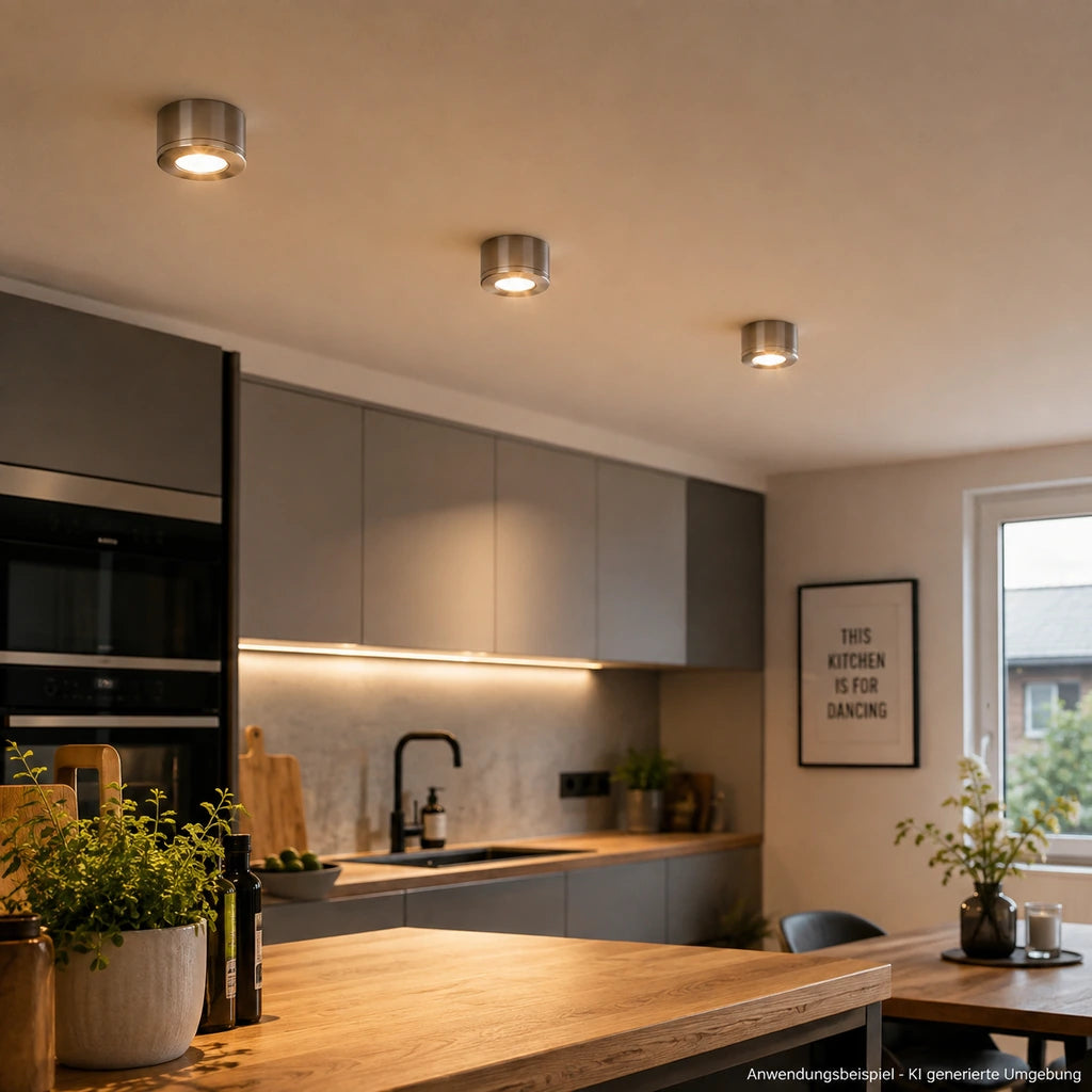 Drei Aluminium LED-Aufbauleuchten an der Decke einer modernen Küche mit warmweißem Licht, Holzarbeitsplatte und grauen Küchenfronten.