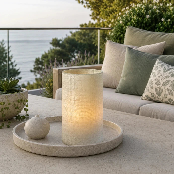 Beige strukturierte Glasvase mit sichtbarem Kerzenlicht auf einem Dekotablett auf einer modernen Terrasse mit grünen Akzenten.