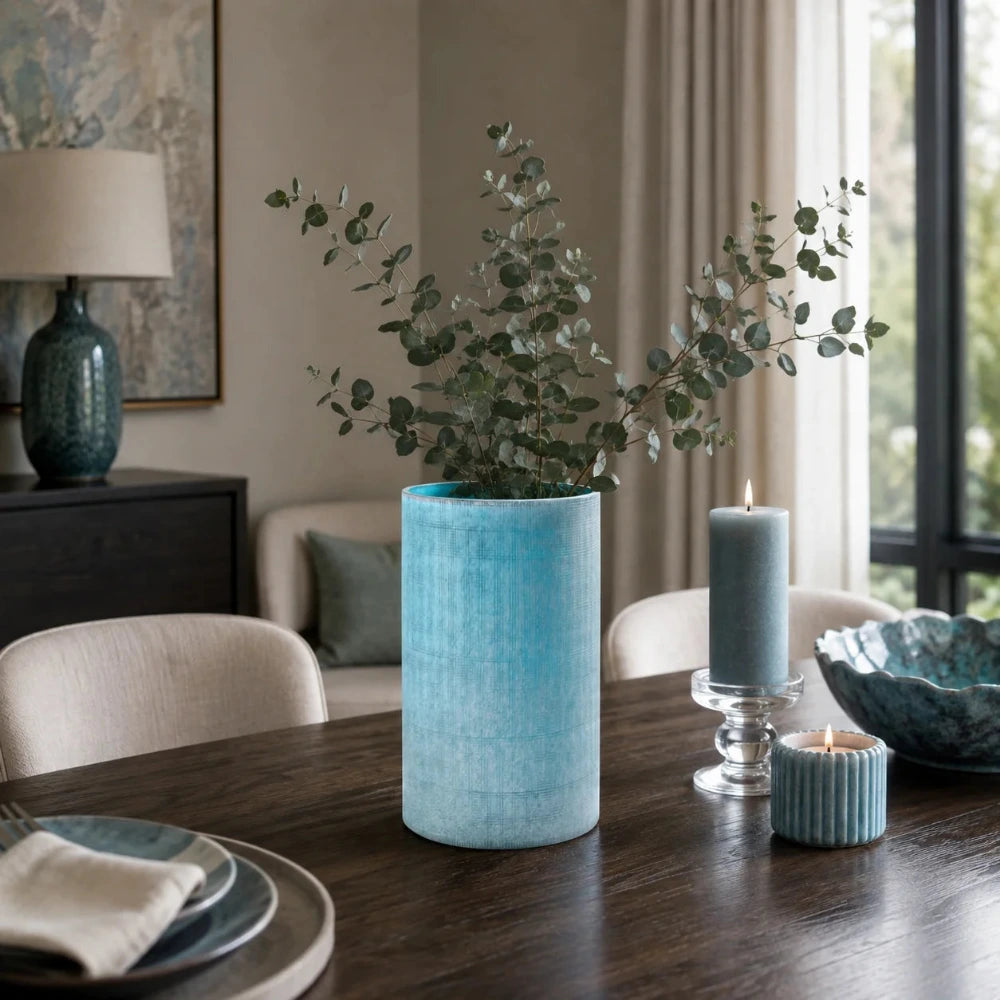 Blaue Glasvase in Vintage-Optik mit grünen Zweigen auf einem modernen Esstisch mit passenden Kerzen