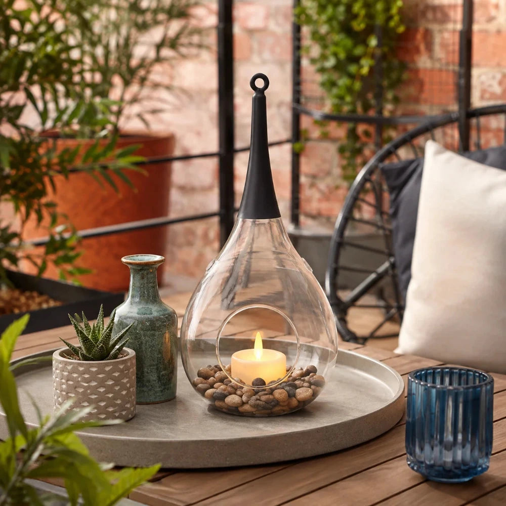 Sompex Taura Hängewindlicht aus klarem mundgeblasenem Glas auf einer Terrasse mit Dekosteinen und LED-Kerze.
